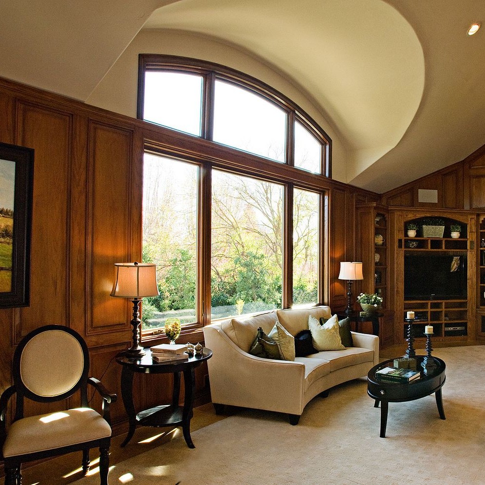 Unequal leg arch windows flank an equal leg arch window. These are atop extremely large picture windows in an opulent wood-paneled den.